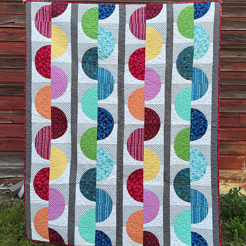 A colorful quilted blanket or small quilt with a geometric pattern of vertical stripes and repeating semicircles in multiple colors, displayed outdoors against a weathered red-brown wooden wall.