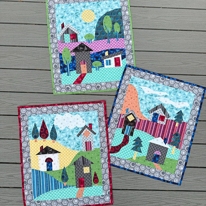 Three small quilted fabric art pieces with whimsical house and landscape designs, arranged on a gray wood plank background.
