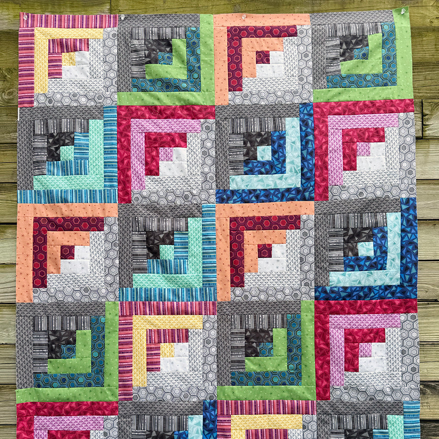 A colorful quilted patchwork blanket with a log cabin pattern, displayed flat against a rustic wooden fence background.