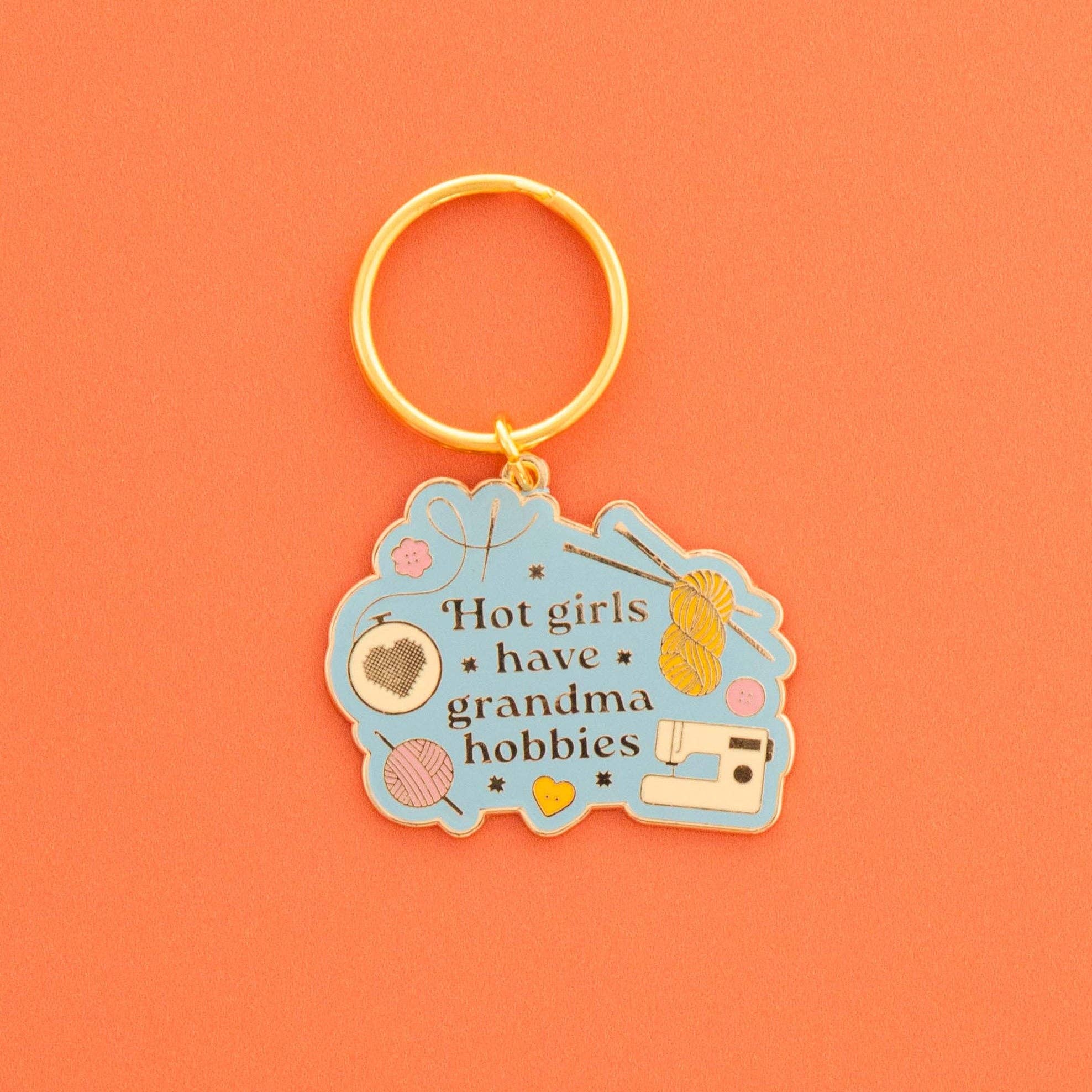 A light blue enamel keychain with a gold metal split ring, featuring the text 'Hot girls have grandma hobbies' and illustrations of knitting needles, yarn balls, a sewing machine, and a heart on a solid orange background.