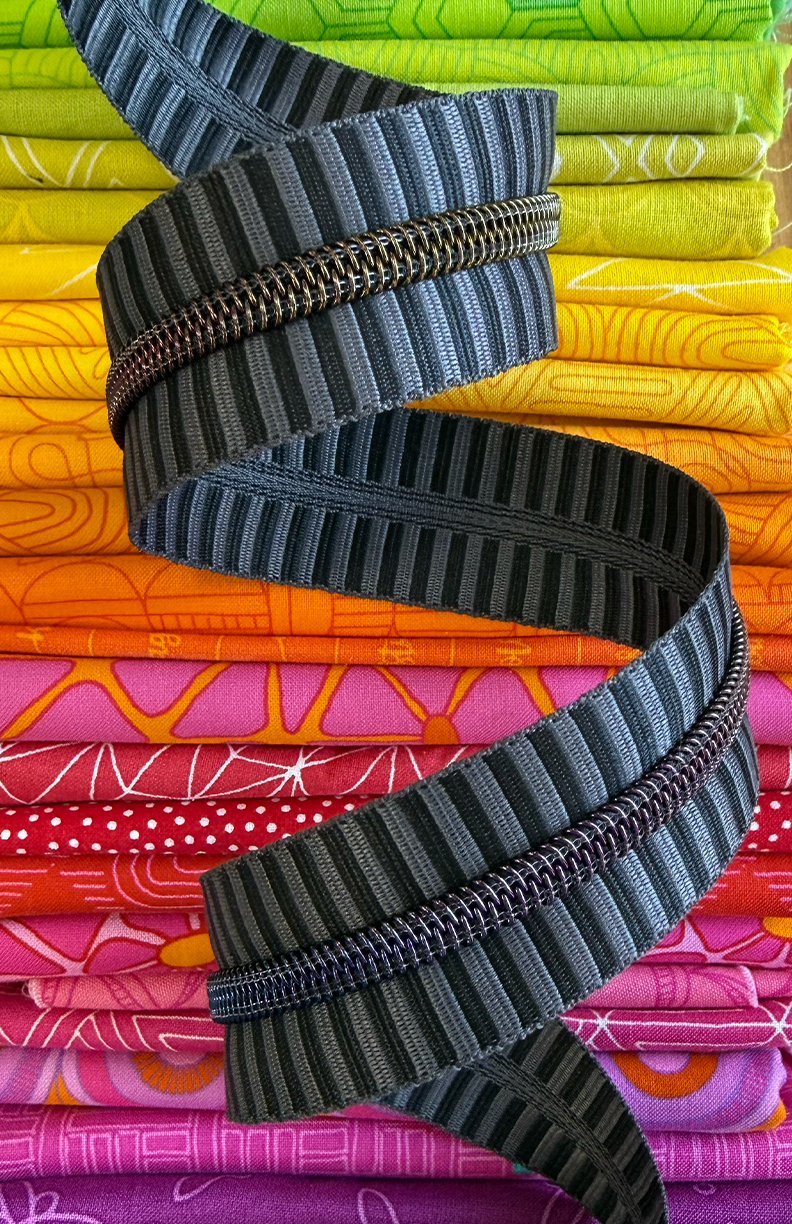 Rainbow zipper rolls with a grey and black striped zipper tape. Photographed on top of beautiful fabric.